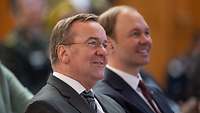 Boris Pistorius und Nils Hilmer sitzen nebeneinander in einem Publikum, beide lächeln.