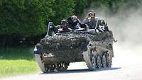 Ein gepanzertes Fahrzeug der Bundeswehr fährt auf einer Straße, zwei Soldaten und eine zivile Person bemannen das Fahrzeug. 